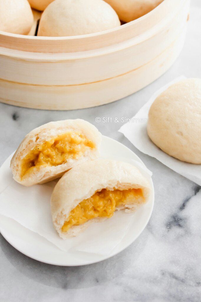 Torn open Custard Buns Lai Wong Bao on white round plate and bamboo steamer.