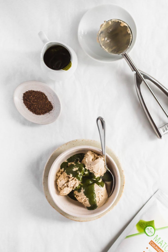 Bowl of hojicha ice cream with matcha sauce drizzled over top, tea packaging, spoon on side on a plate, tea powder in small dish, ice cream scoop.