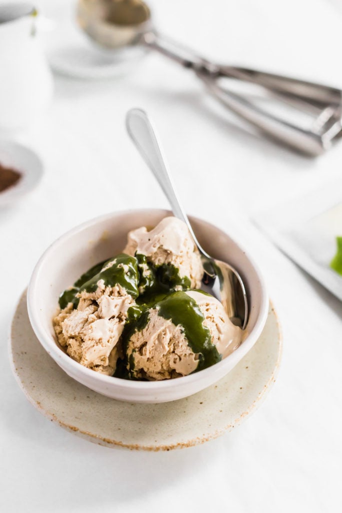 Bowl of hojicha ice cream with matcha sauce drizzled over top, spoon on side on a plate.