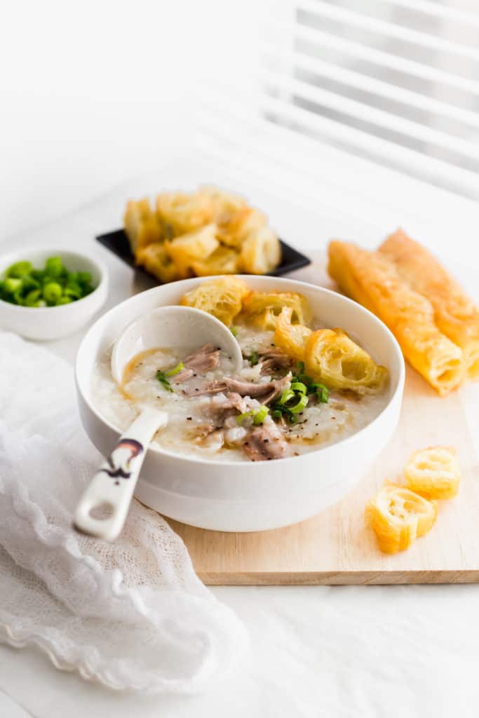 Rice congee with turkey and Chinese donut in white bowl with ladle on wooden board, donut in background.