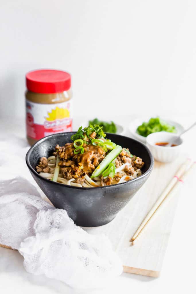 Noodles with pork topping in a black bowl, chopsticks on the side, SunButter jar in background.
