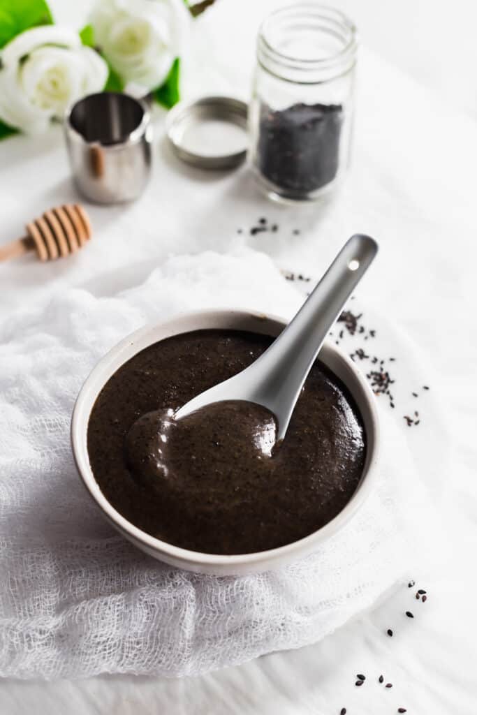 Close up of black sesame soup with a spoon in a white bowl on cheesecloth and marble trivet, black sesame seeds scattered around.