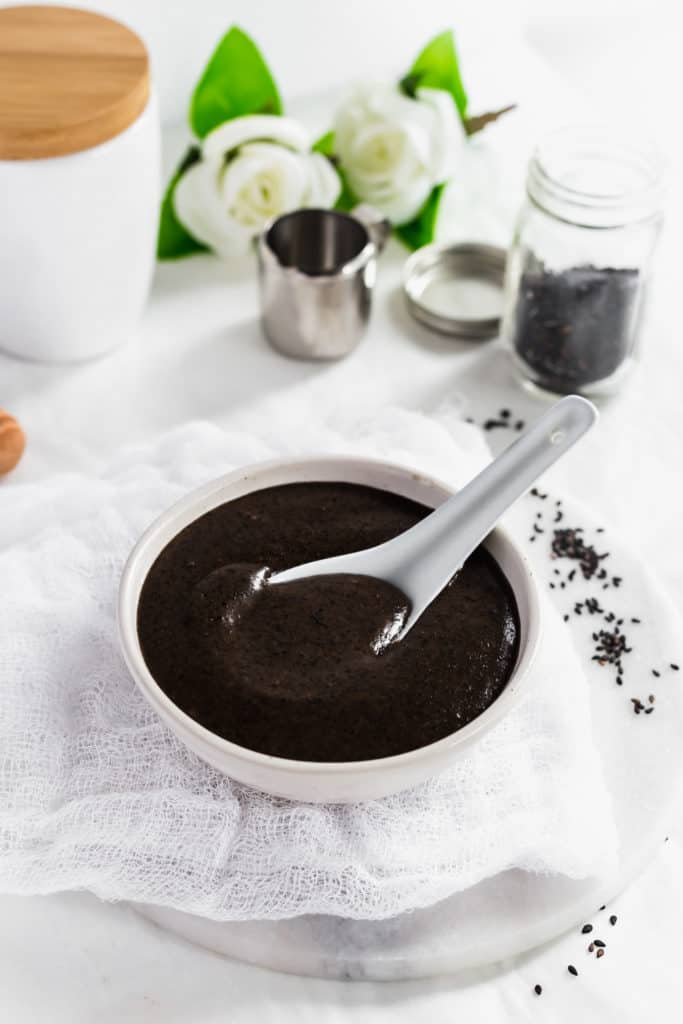Black sesame soup with a spoon in a white bowl on cheesecloth and marble trivet, black sesame seeds scattered around.