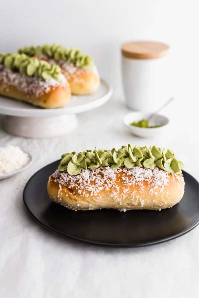 A close up of coconut bun with matcha cream on a black round plate, matcha in small dish with spoon, buns in background