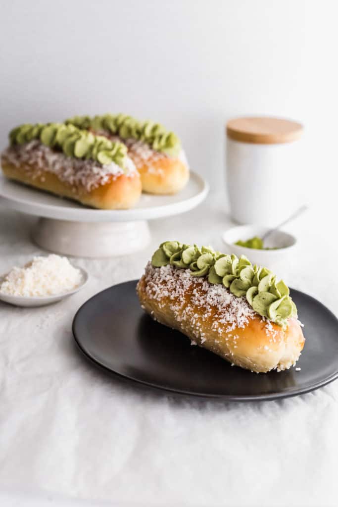 A close up of coconut bun with matcha cream on a black round plate, matcha in small dish with spoon, buns in background