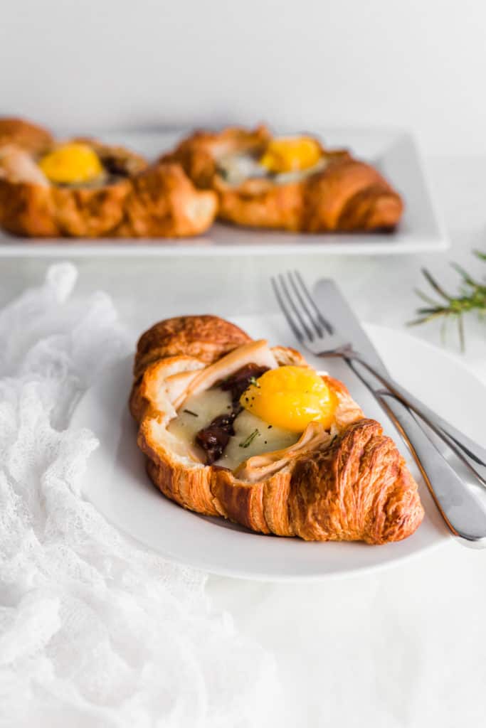 A turkey and egg croissant on a round plate with fork and knife on side, croissants in background.