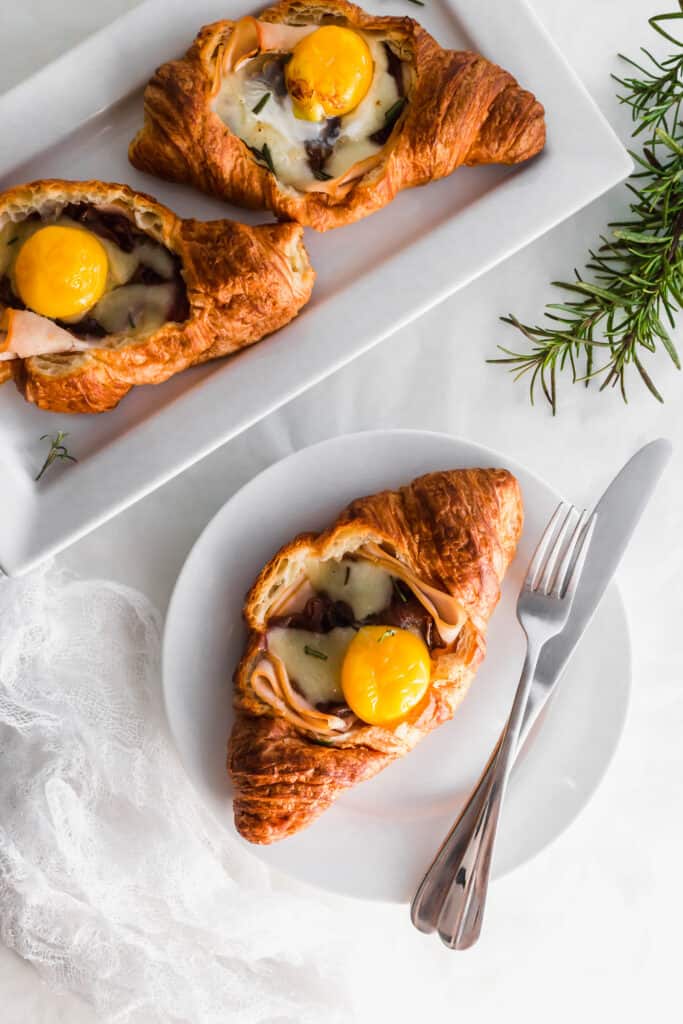 A turkey and egg croissant on a round plate with fork and knife on side, croissants on rectangular plate.