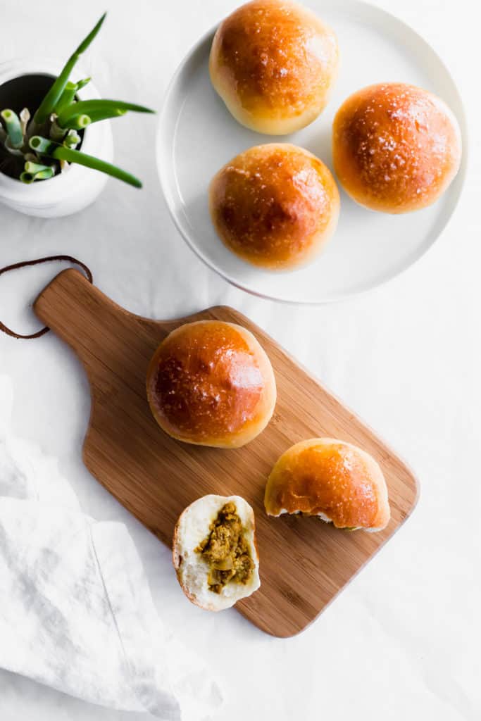 A torn curried turkey bun revealing the interior on a wooden board, buns on a circular white plate.