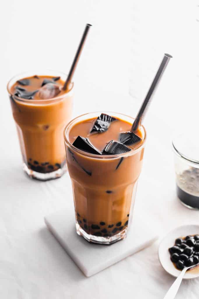 Glasses of HK milk tea with boba pearls and grass jelly, straw, on a white marble coaster, small plate of boba pearls on the side.