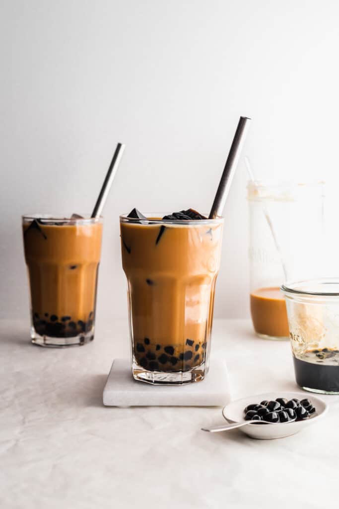 Glasses of HK milk tea with boba pearls, straw, on a white marble coaster, small plate of boba pearls on the side.