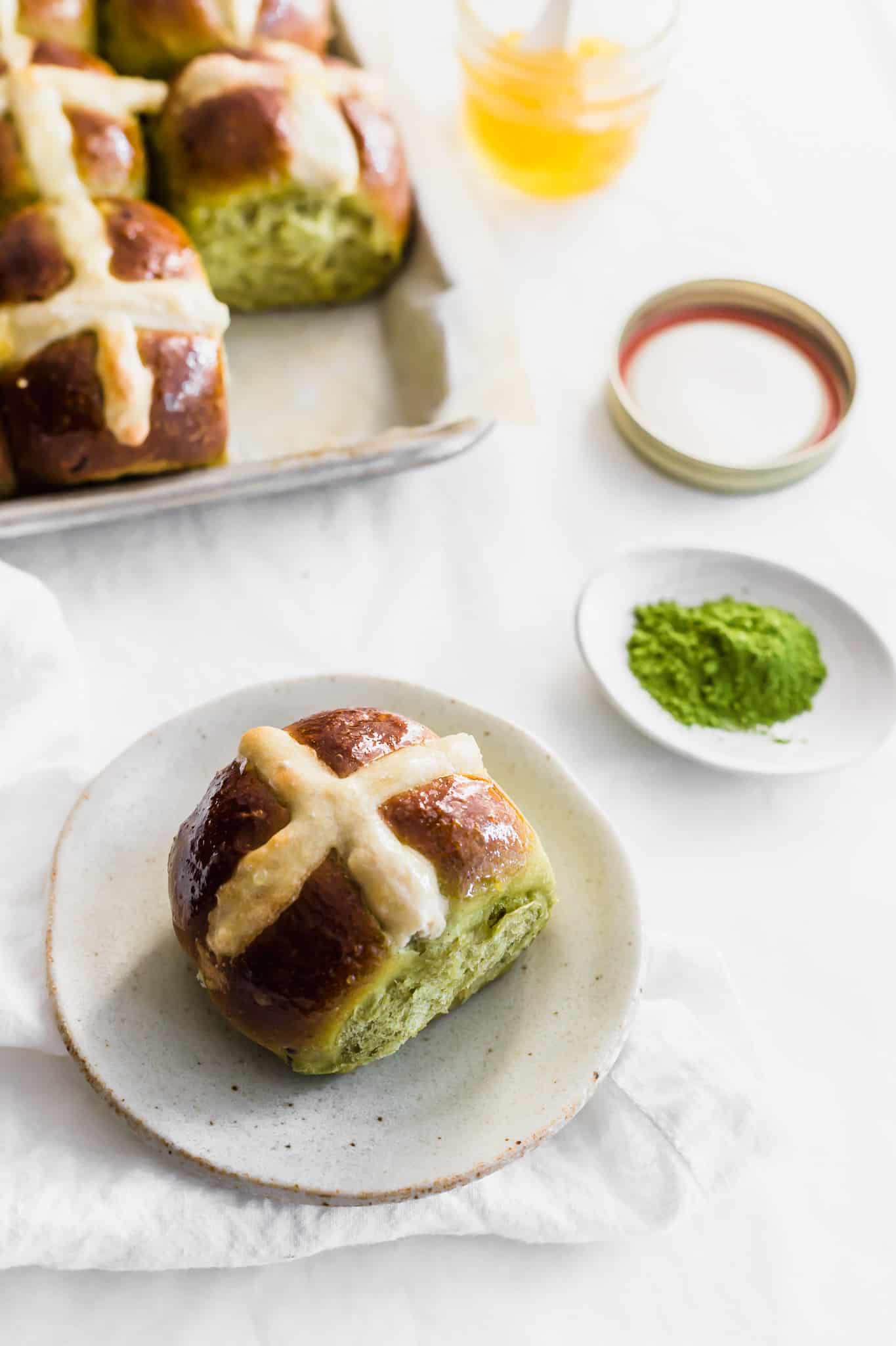 A close up of matcha yuzu hot cross bun on a small plate on white linen, matcha powder in dish on the side, yuzu jam in jar, buns in a tray in background.