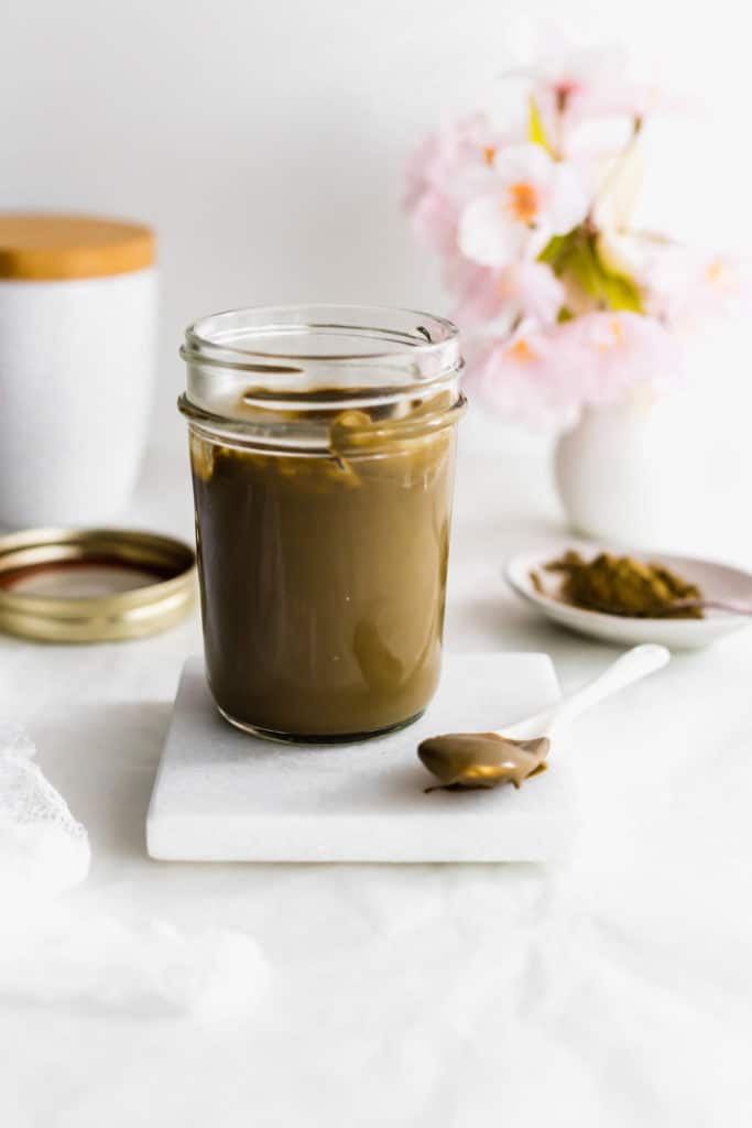 Jar of Hojicha Roasted Green Tea Milk Jam on marble coaster, spoon on side.