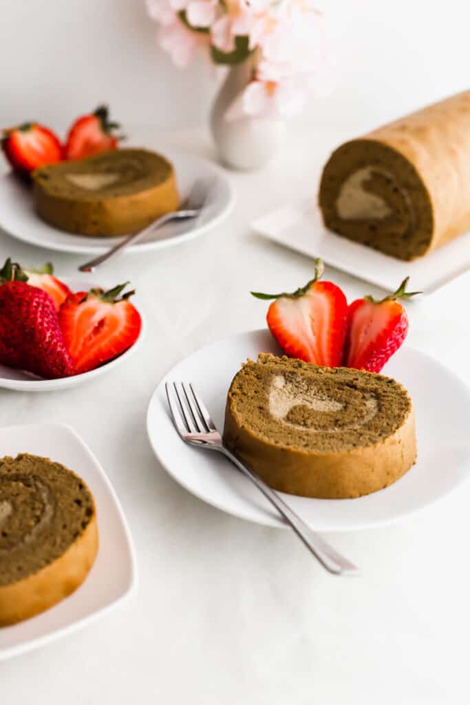 A slice of hojicha roll cake on a round plate with fork and cut strawberries, strawberries and uncut cake in the background.
