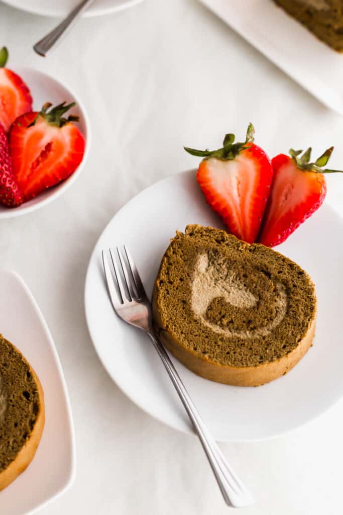 A slice of hojicha roll cake on a round plate with fork and cut strawberries.