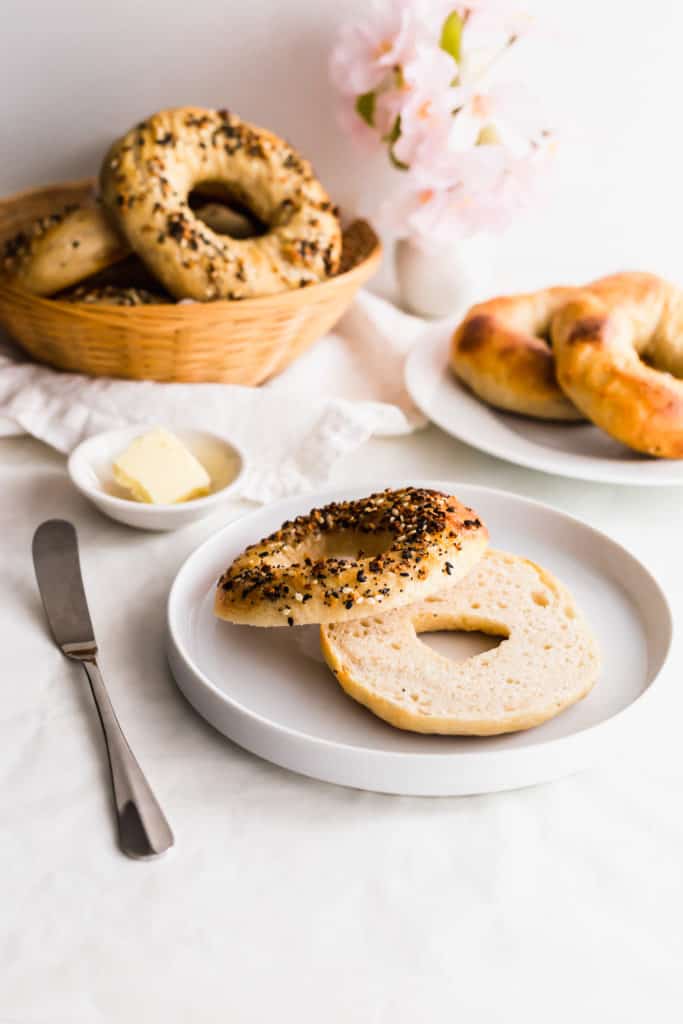 A sliced open sourdough bagel on a white plate with knife on side, butter in small dish, bagels in basket and on a plate in background.