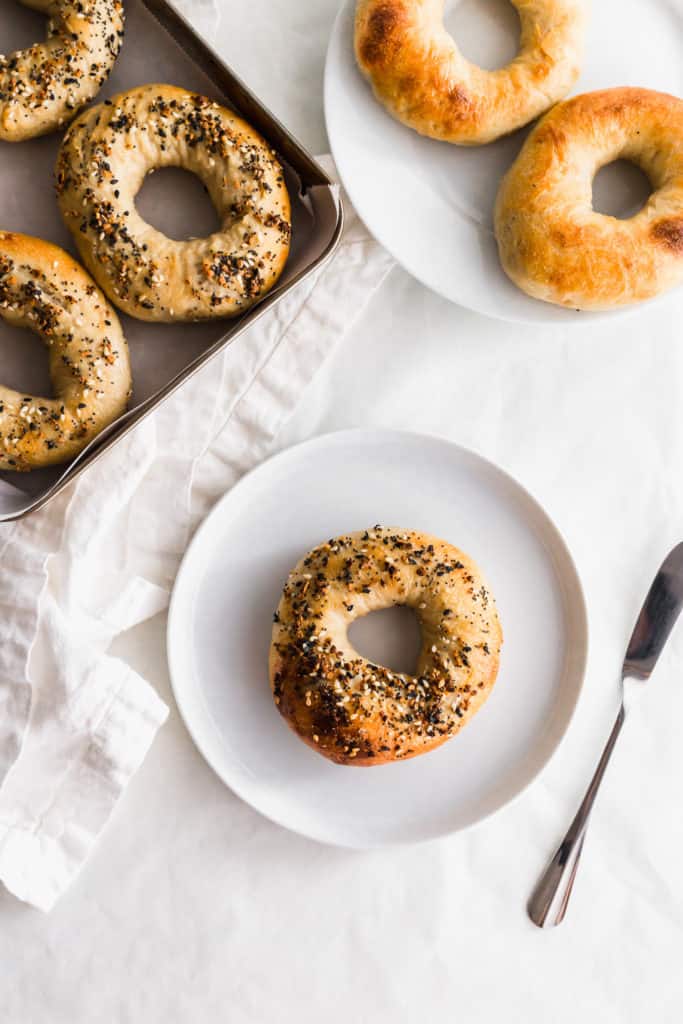 A sourdough bagel on a white plate with knife to the side, bagels on a tray and plate.