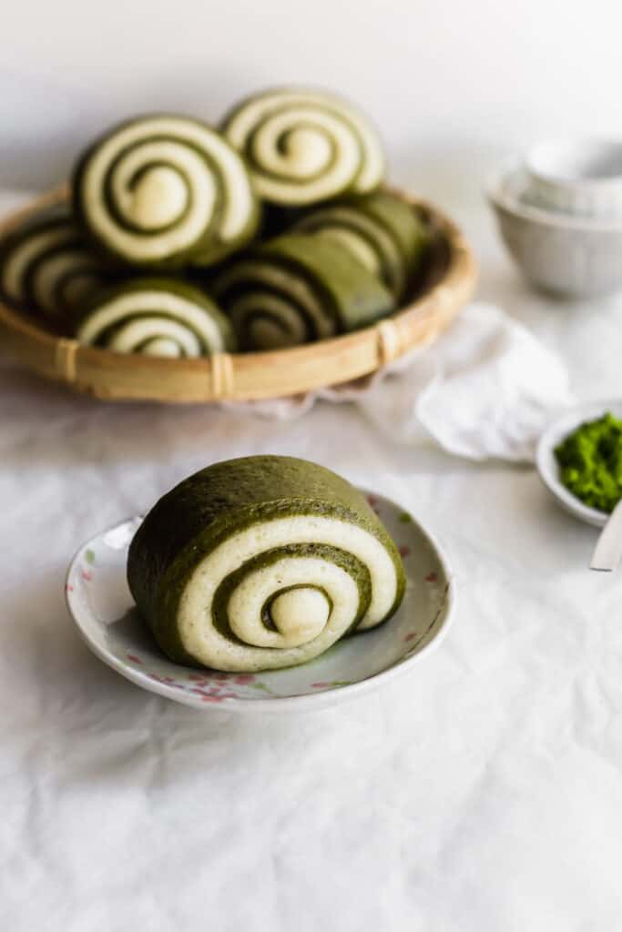 A swirled green and white matcha mantou bun on small plate with buns in the background.