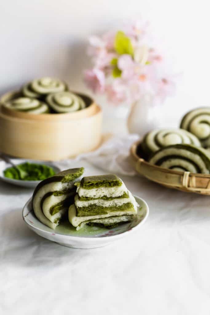 Torn green and white matcha mantou bun revealing interior on small plate with matcha tea, more buns in bamboo steamers in background.