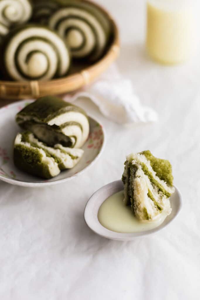 A close up of torn green and white mantou bun dipped in condensed milk on a small plate, ripped bun on a plate in background.