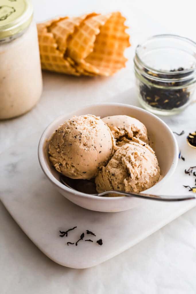 Scoops of ice cream in a bowl with a spoon on marble with earl grey tea in a jar scattered around, waffles cones in background.