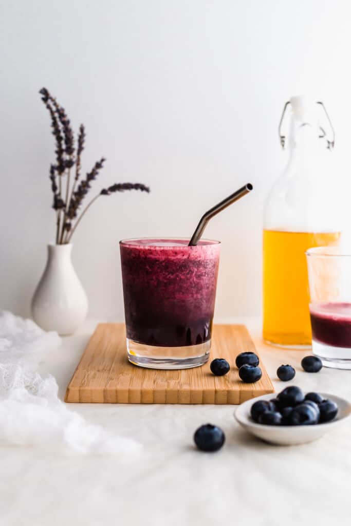 A glass of lavender blueberry jasmine tea on a wooden board with blueberries scattered around, yellow jasmine tea and lavender flowers in the background.