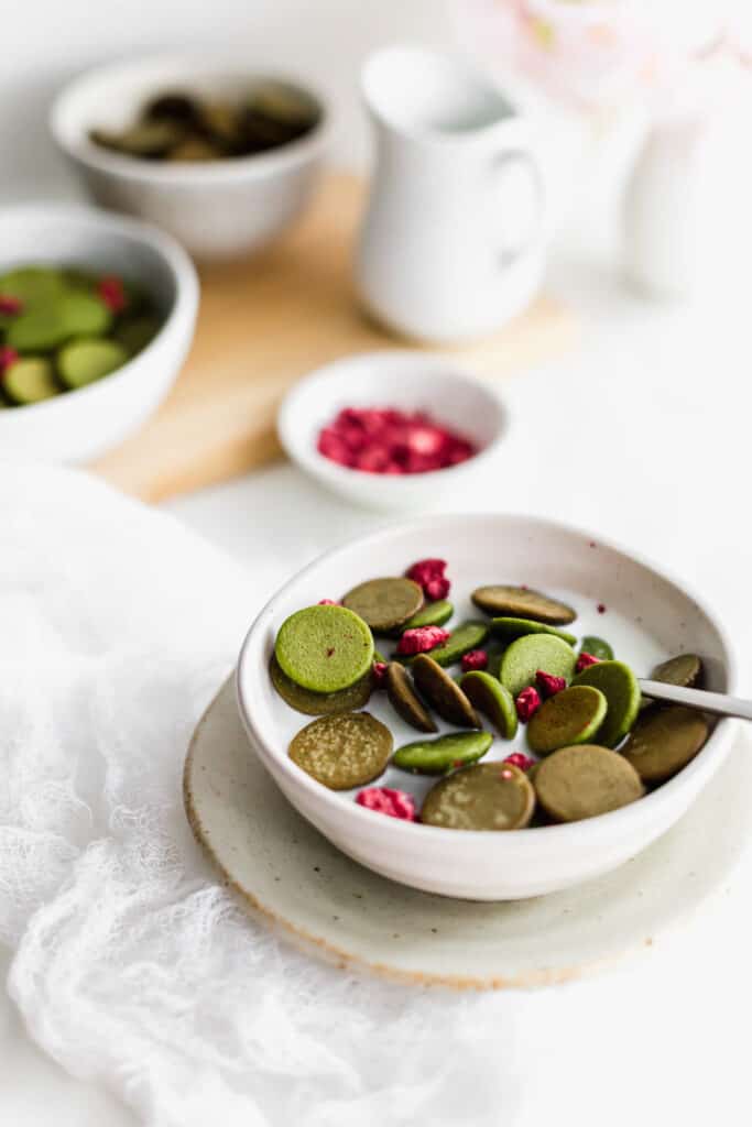 A spoonful of mini hojicha and matcha pancakes with dried raspberries in milk in a bowl on a small plate, jug of milk, raspberries and mini pancakes in background.