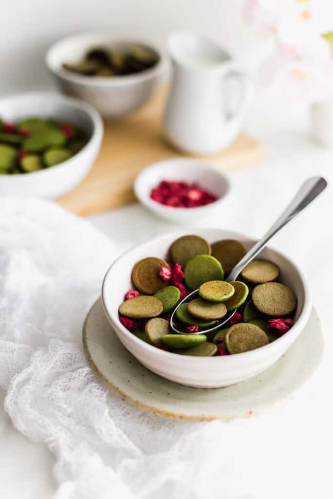A spoonful of mini hojicha and matcha pancakes with dried raspberries in a bowl on a small plate, jug of milk, raspberries and mini pancakes in background.