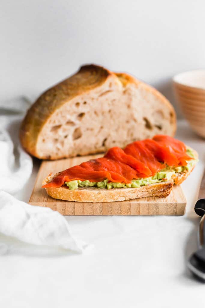 A slice of sourdough bread topped with avocado and salmon on wooden board with sourdough bread in background.