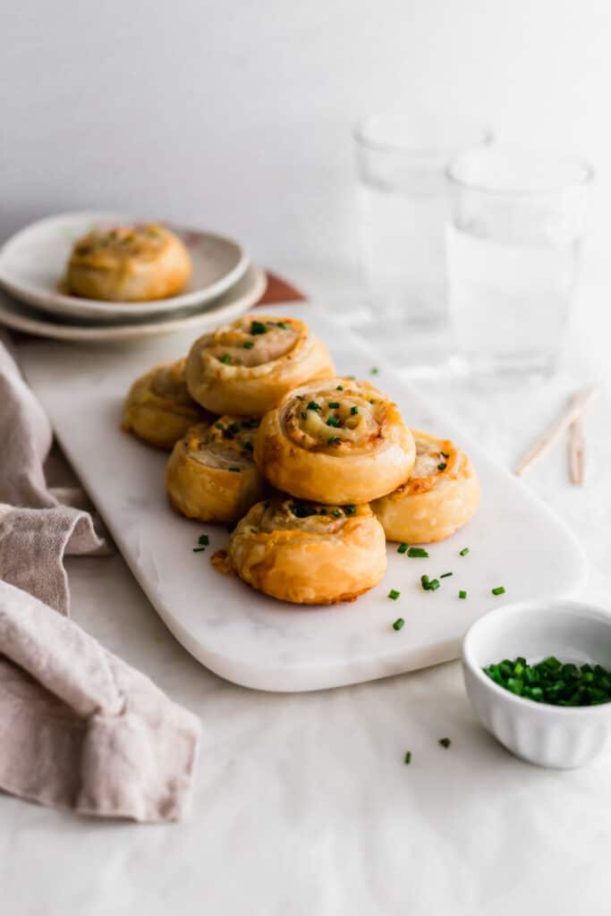 A stack of ham and cheese puff pastry scrolls topped with chives on marble with water glasses in the background and bowl of chives in the foreground..