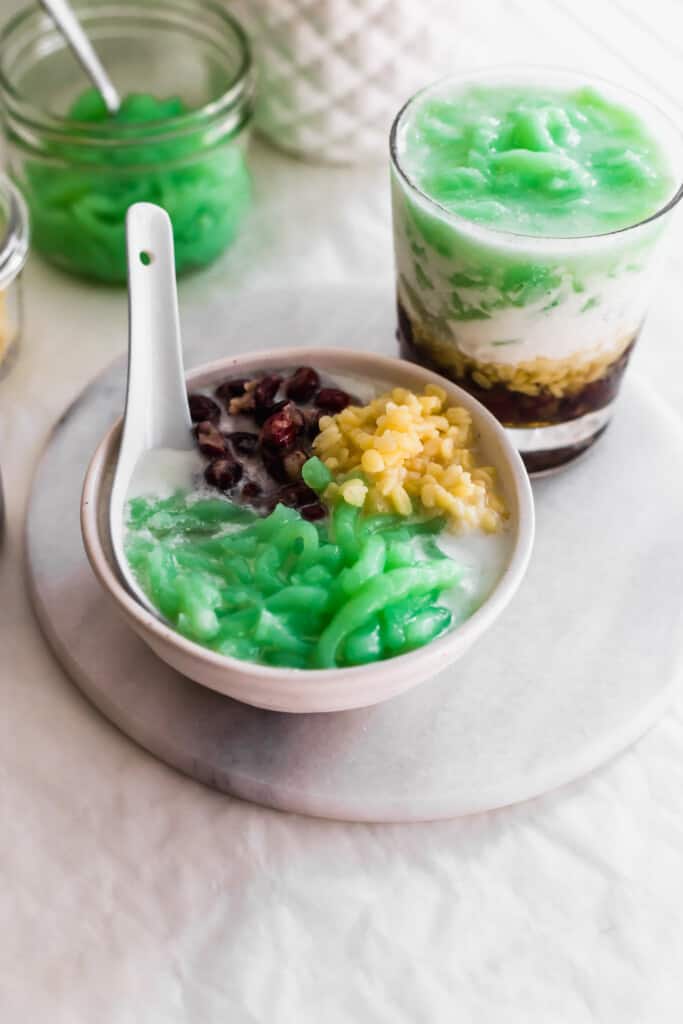 A bowl of cendol green jellies, red bean, yellow mung beans in coconut soup with spoon, glass of layered cendol behind..