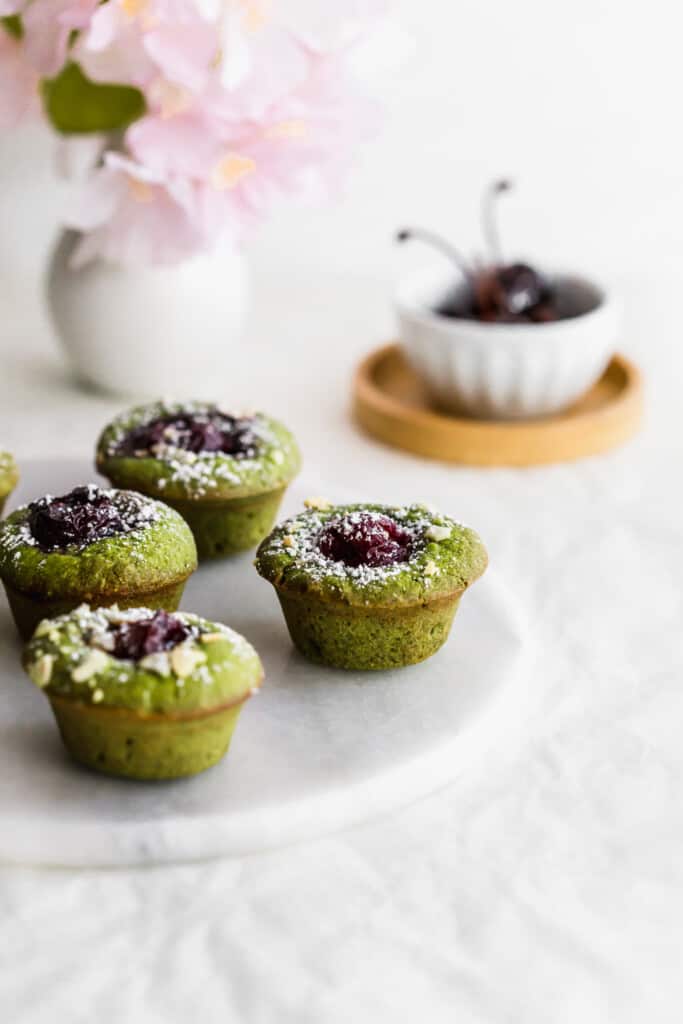 Green matcha and cherry financiers on a round marble trivet with a bowl of cherries in the background.