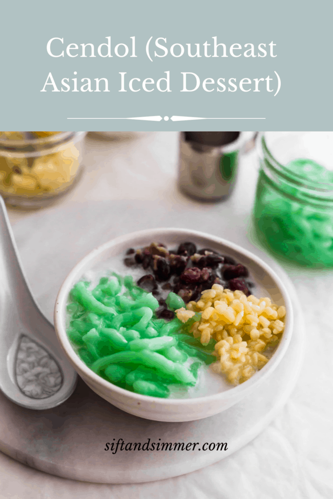 A bowl of cendol green jellies, red bean, yellow mung beans in coconut soup with a spoon on the side, with text overlay. 