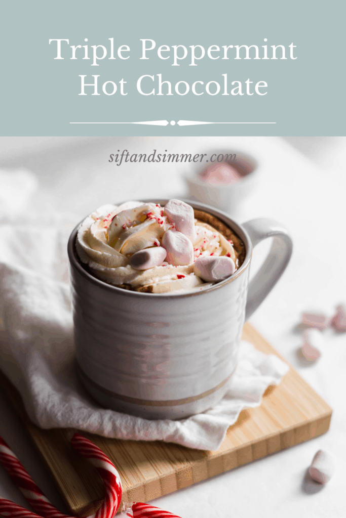 Close up mug of hot chocolate with whipped cream and pink marshmallows on linen on wooden board, candy cane on side, marshmallows scattered around.