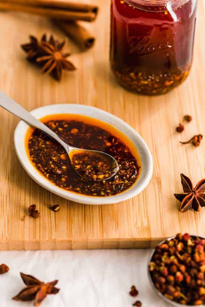 Dish of chili oil with spoon, spices on wooden board.