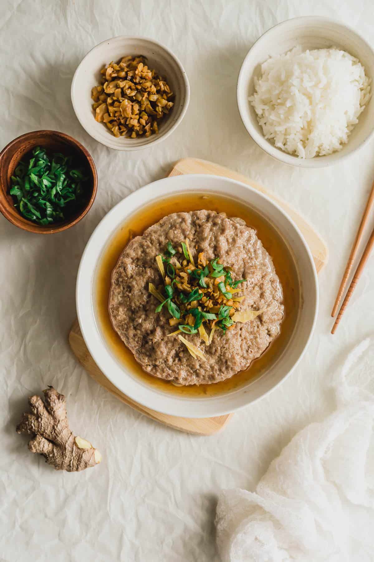 Chinese steamed pork patty in a white plate on wooden board, with a bowl of rice and chopsticks beside it.