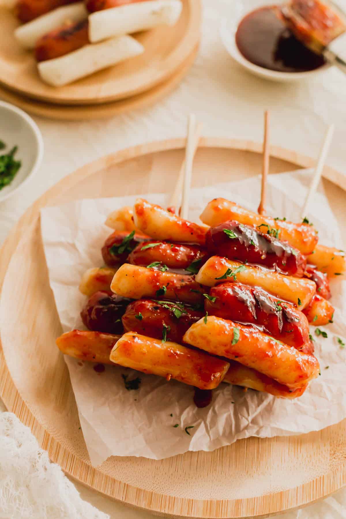 Sotteok skewered rice cakes and sausages on a piece of white parchment paper on wooden round plate.