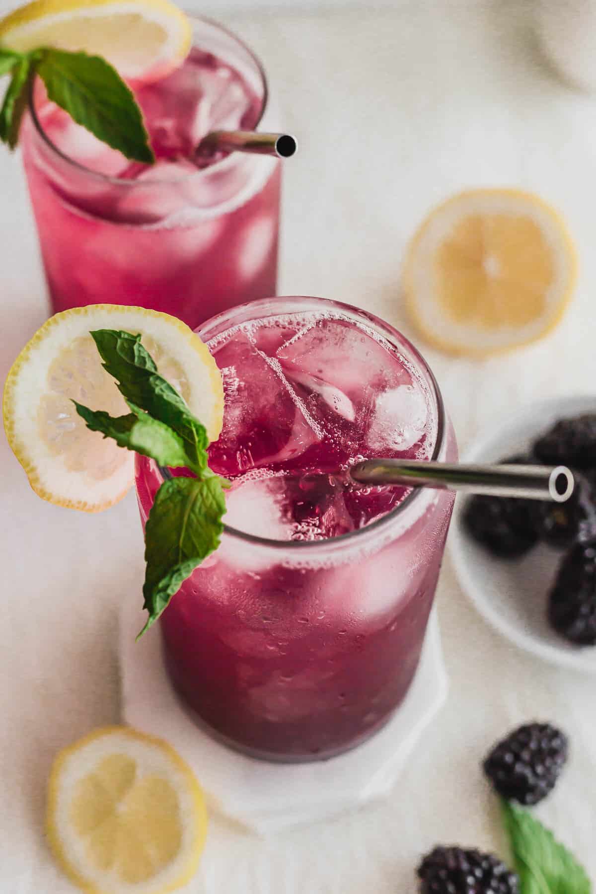 Sparkling blackberry yuzu drinks in glass with metal straws on stacked white coasters.