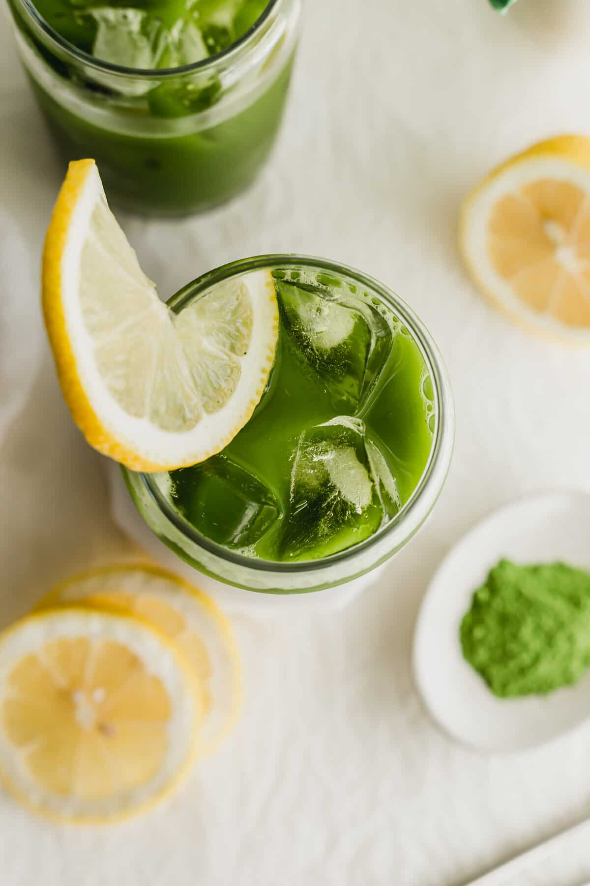 Glasses of matcha tea lemonade with lemon on white coaster.