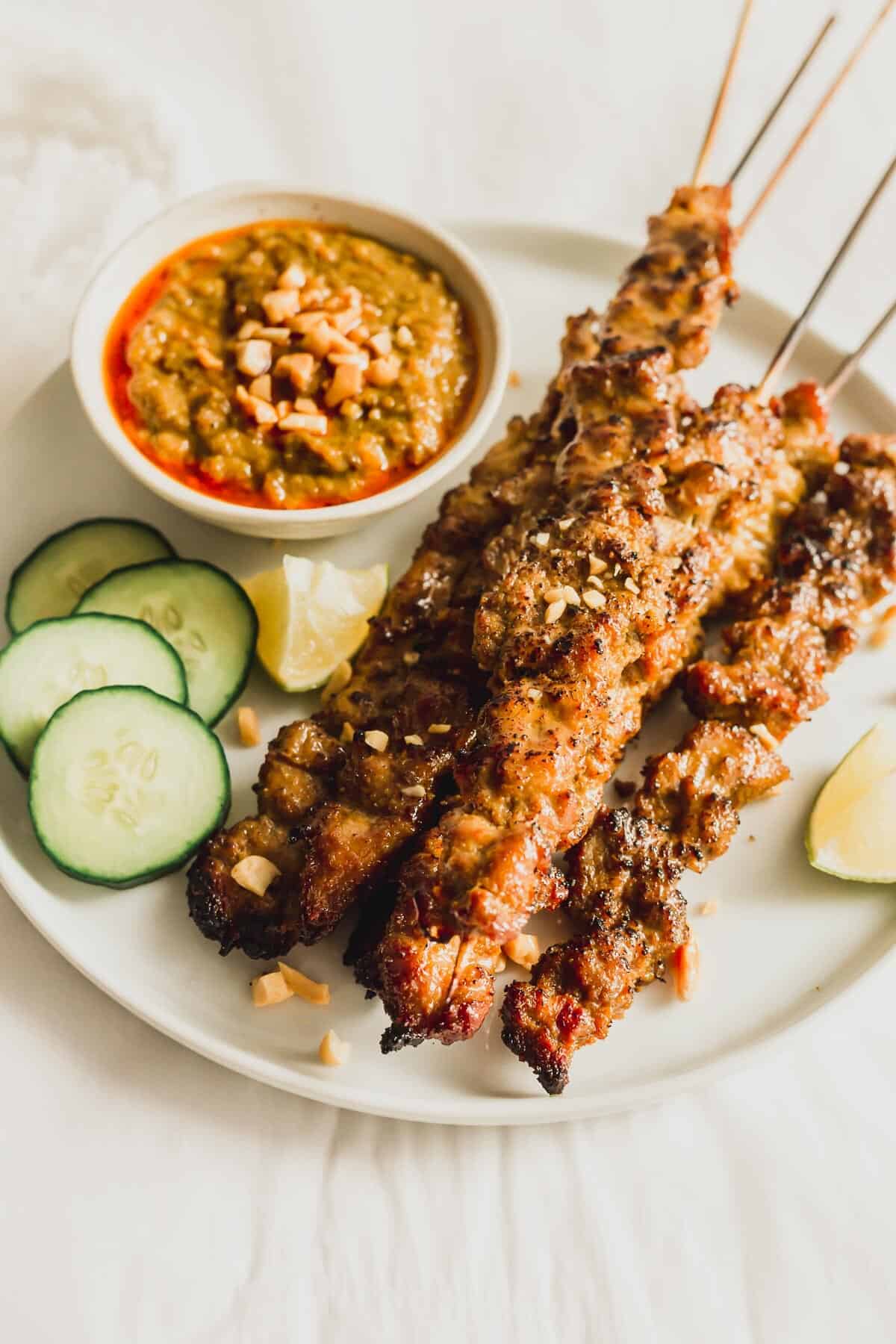 Pork satay skewers on a white round plate with peanut sauce on the side.