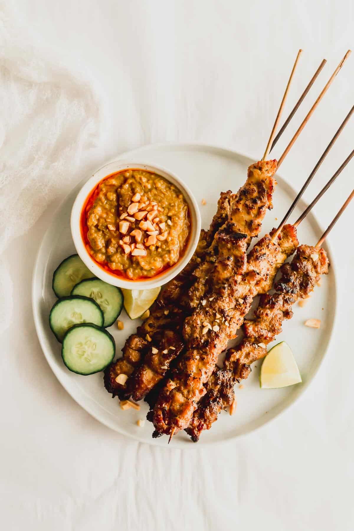 Pork satay skewers on a white round plate with peanut sauce on the side.