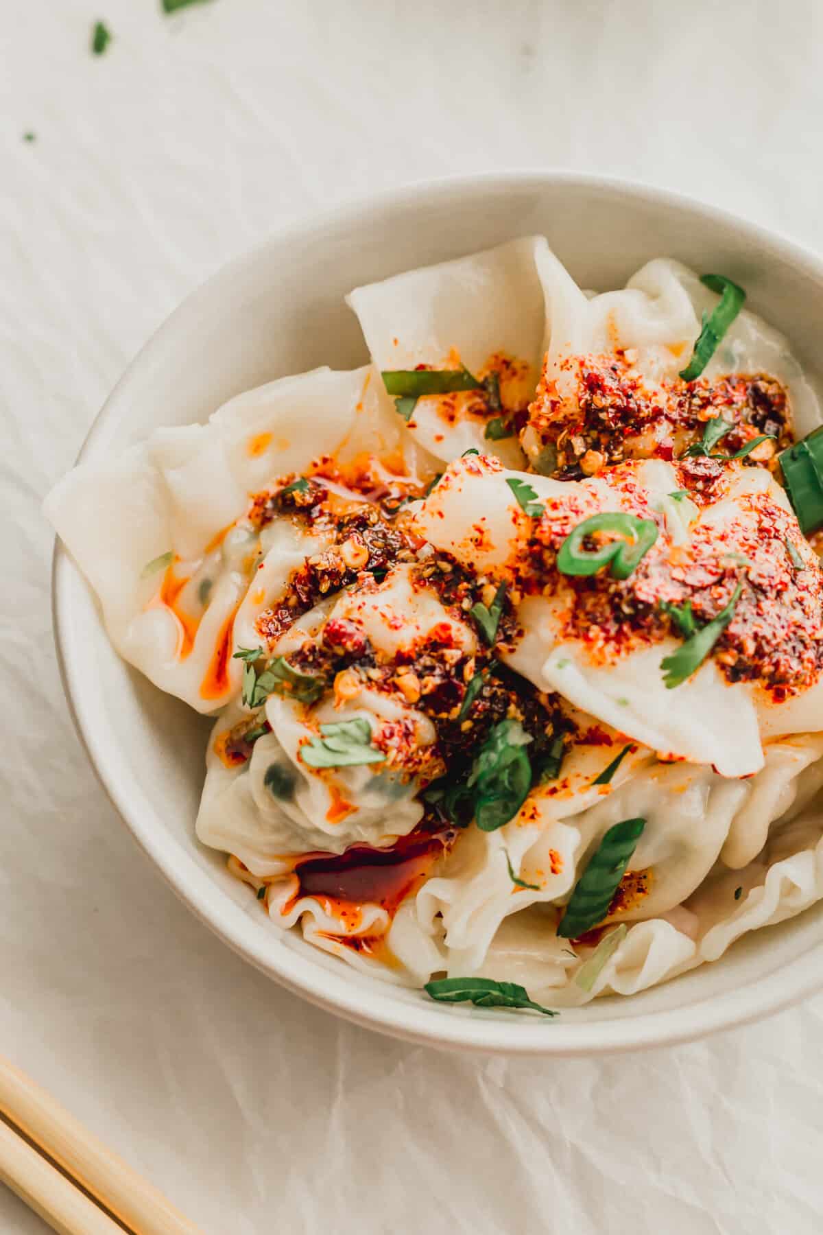 A white ceramic bowl filled with wonton dumplings drizzled with chili oil.