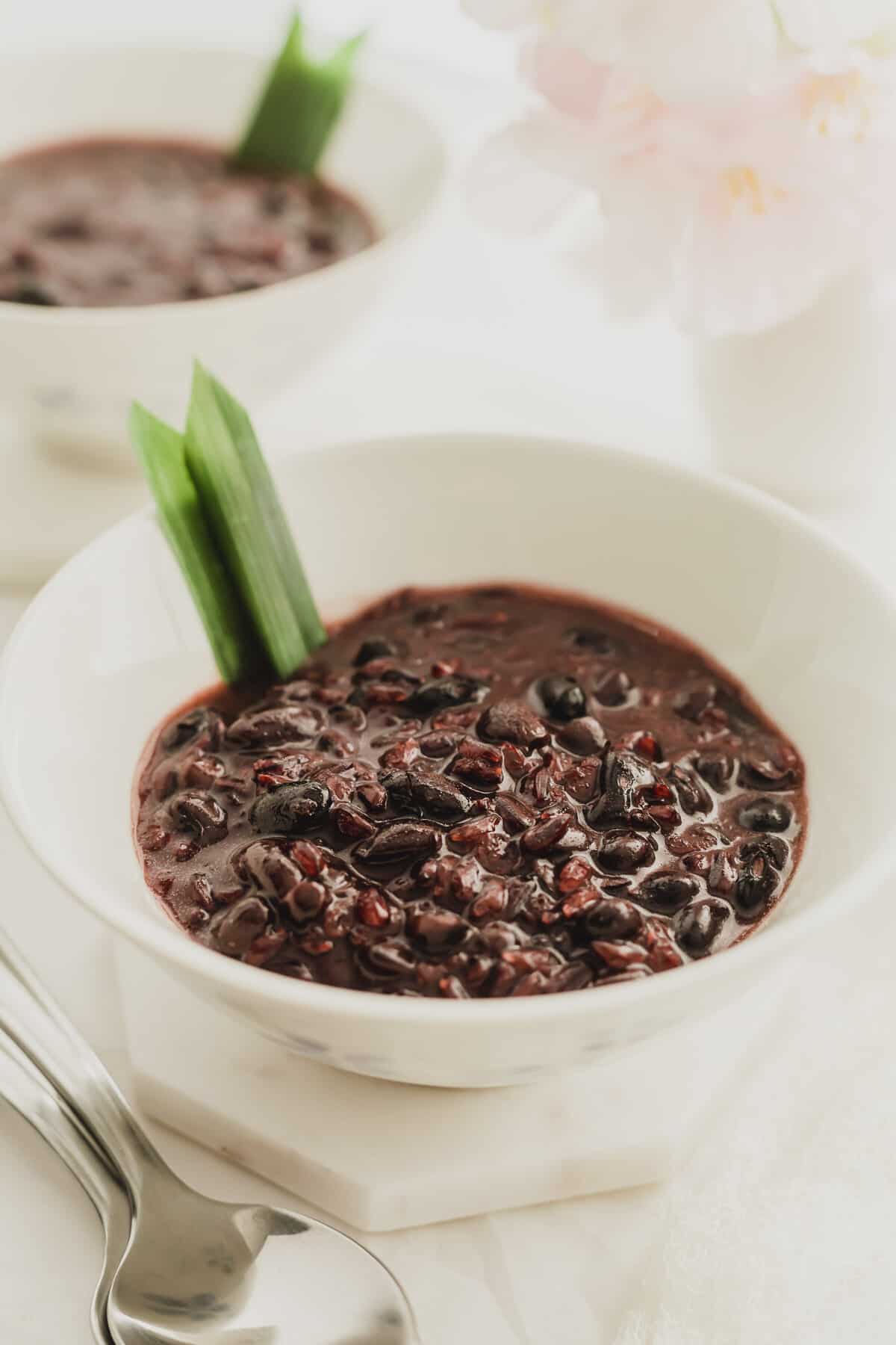 A white bowl filled with black glutinous rice with coconut cream and a pandan leaf.