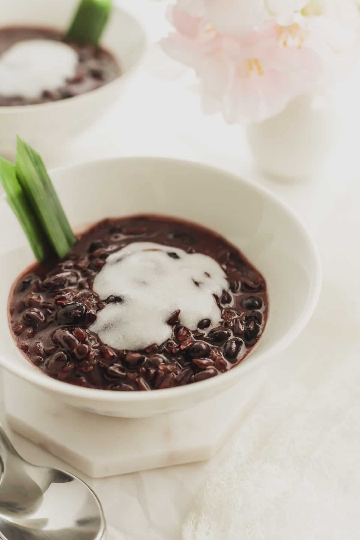 A white bowl filled with black glutinous rice with coconut cream and a pandan leaf.