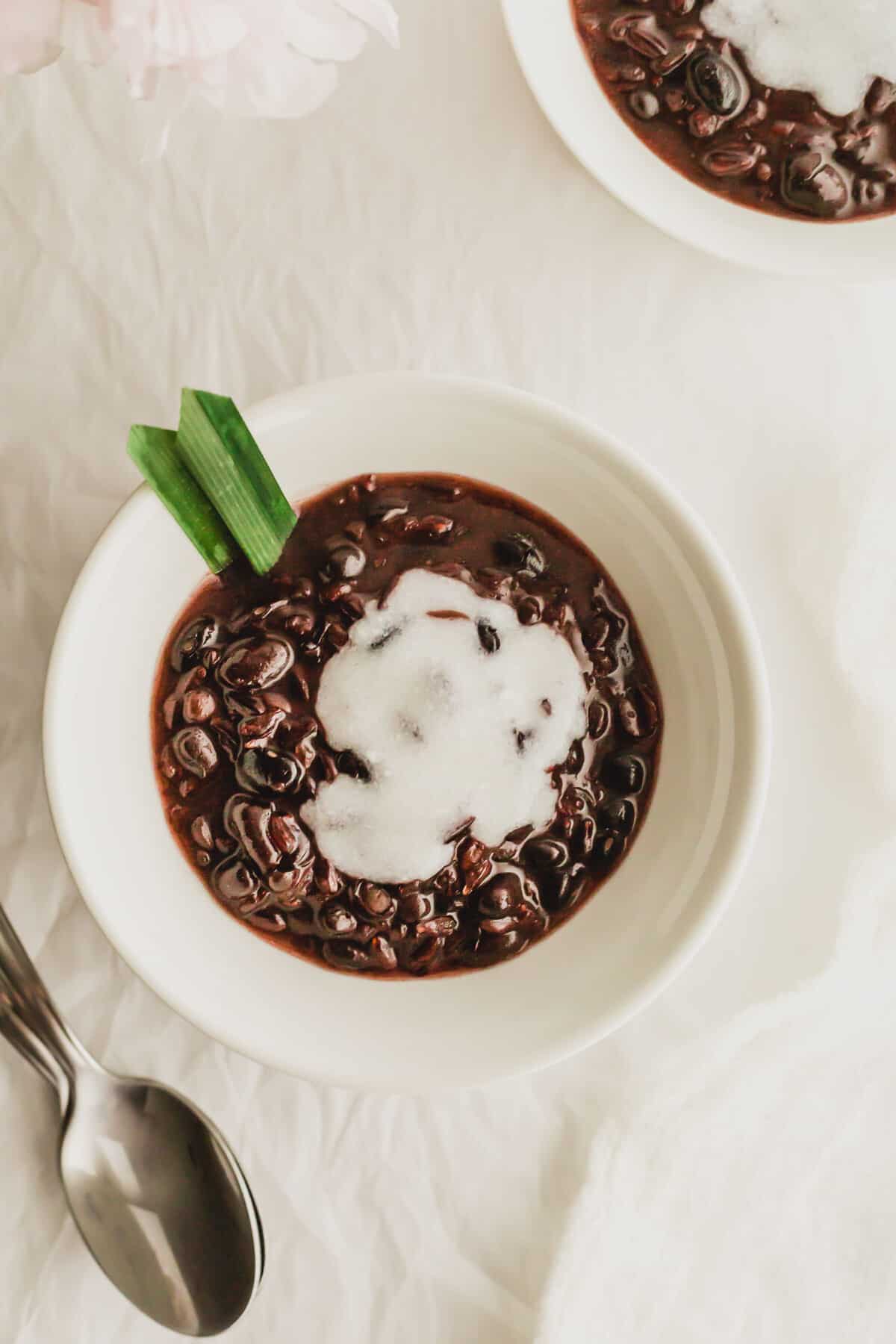 A white bowl filled with black glutinous rice with coconut cream and a pandan leaf.