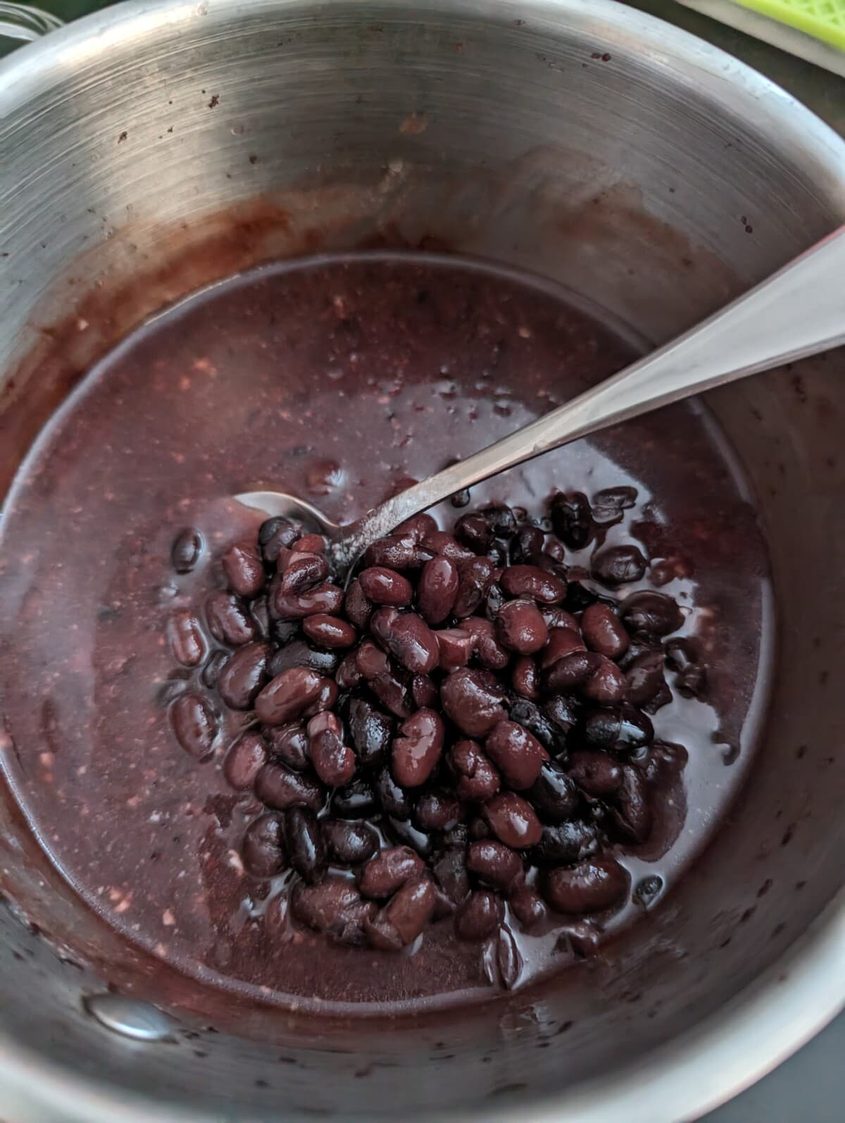 Black beans added to a metal saucepan with cooked black glutinous rice, with a spoon.