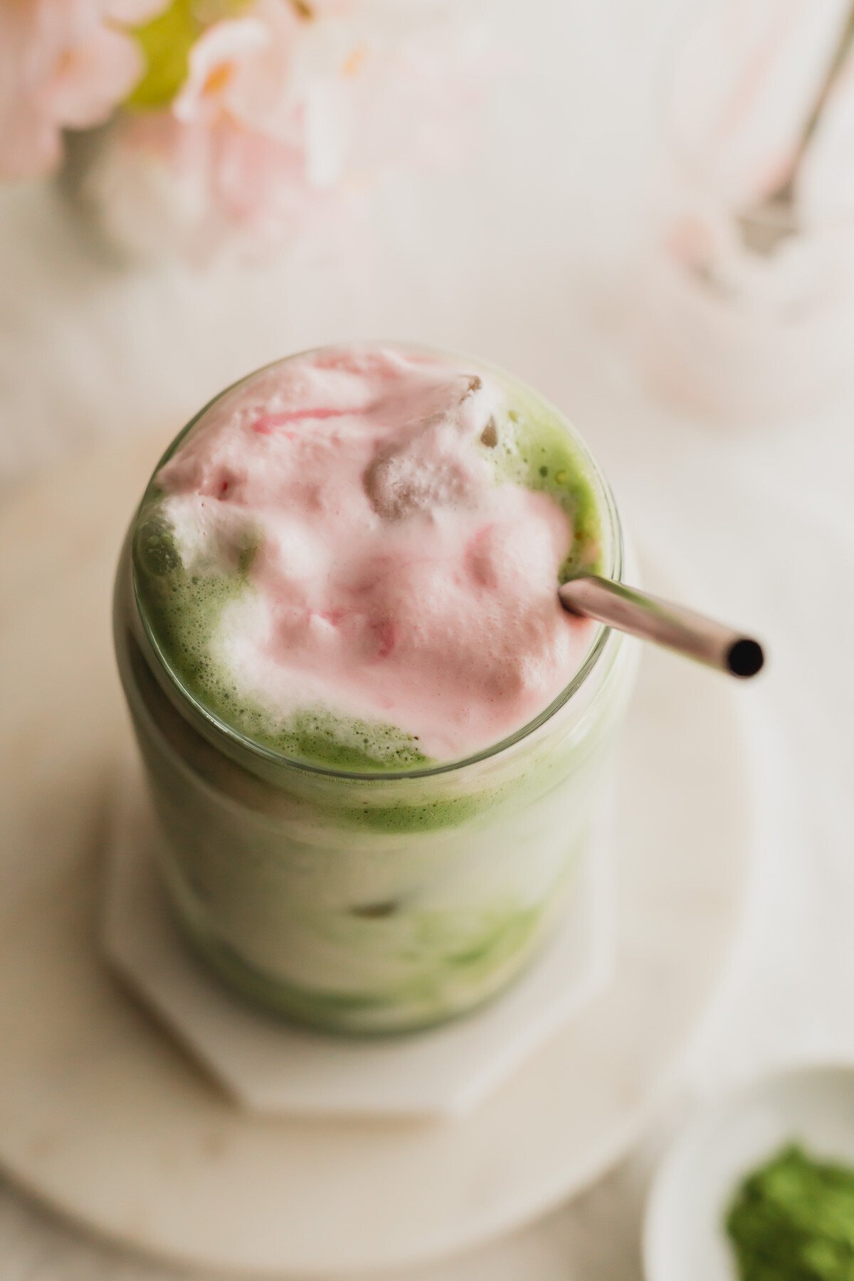 A close up of a glass of sakura matcha latte, with stainless steel straw, on white marble coasters.