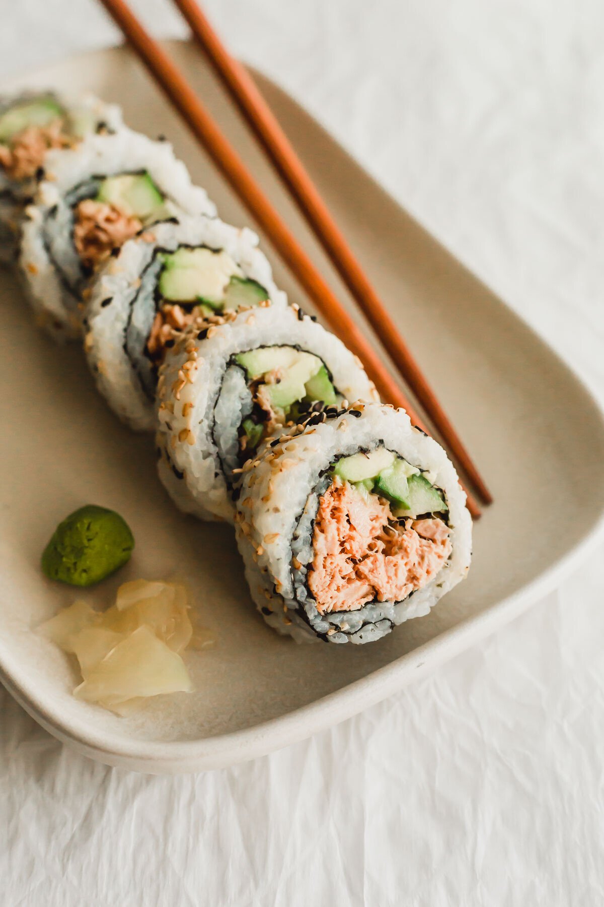 Spicy tuna sushi rolls lined up on a beige rectangular plate with wooden chopsticks on the side.
