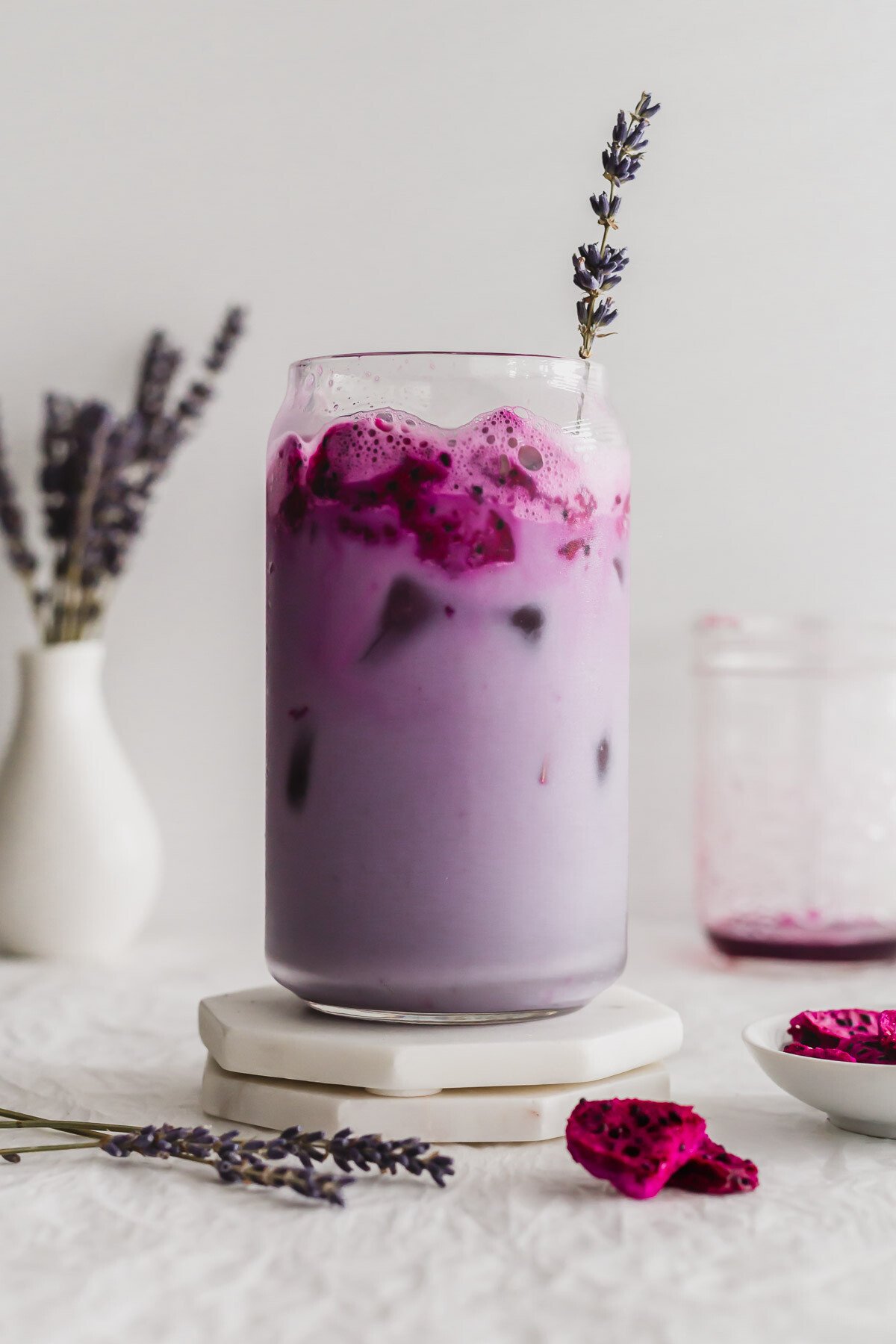 Lavender oat milk with dragon fruit in a glass with lavender sprig, on white marble coasters.
