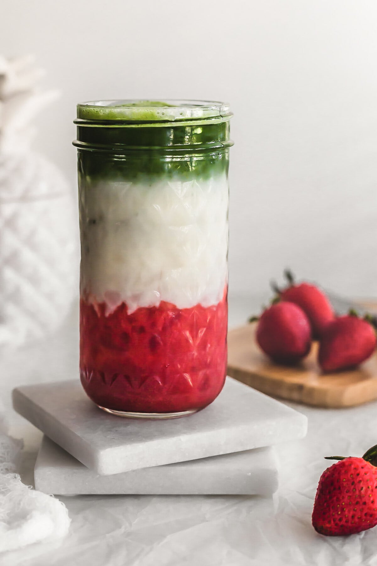 Layered matcha strawberry milk in glass jar on white coasters.