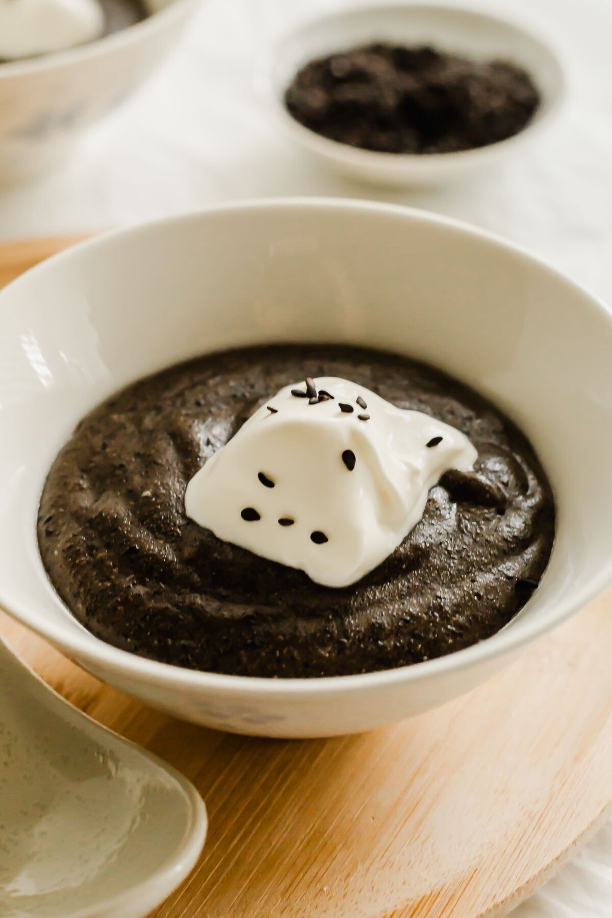 Close up of Black sesame paste in a white bowl, with a dollop of whipped cream and black sesame seeds.